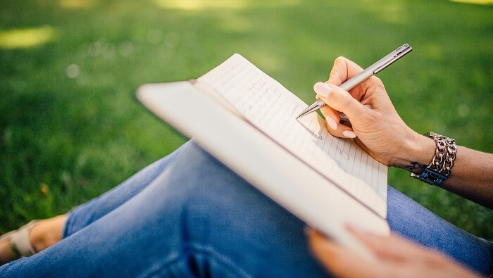 gratitude exercises concept - woman sitting on grass writing in journal